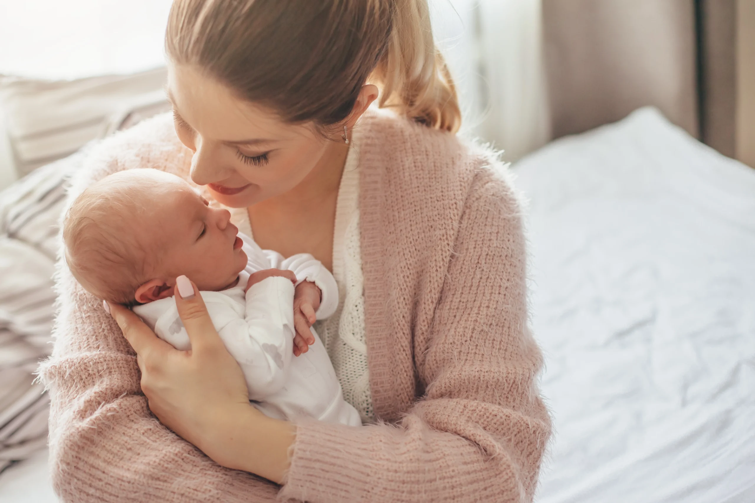 Jak cesarskie cięcie wpływa na mikrobiom niemowląt? Młoda mama w różowym swetrze przytula noworodka.